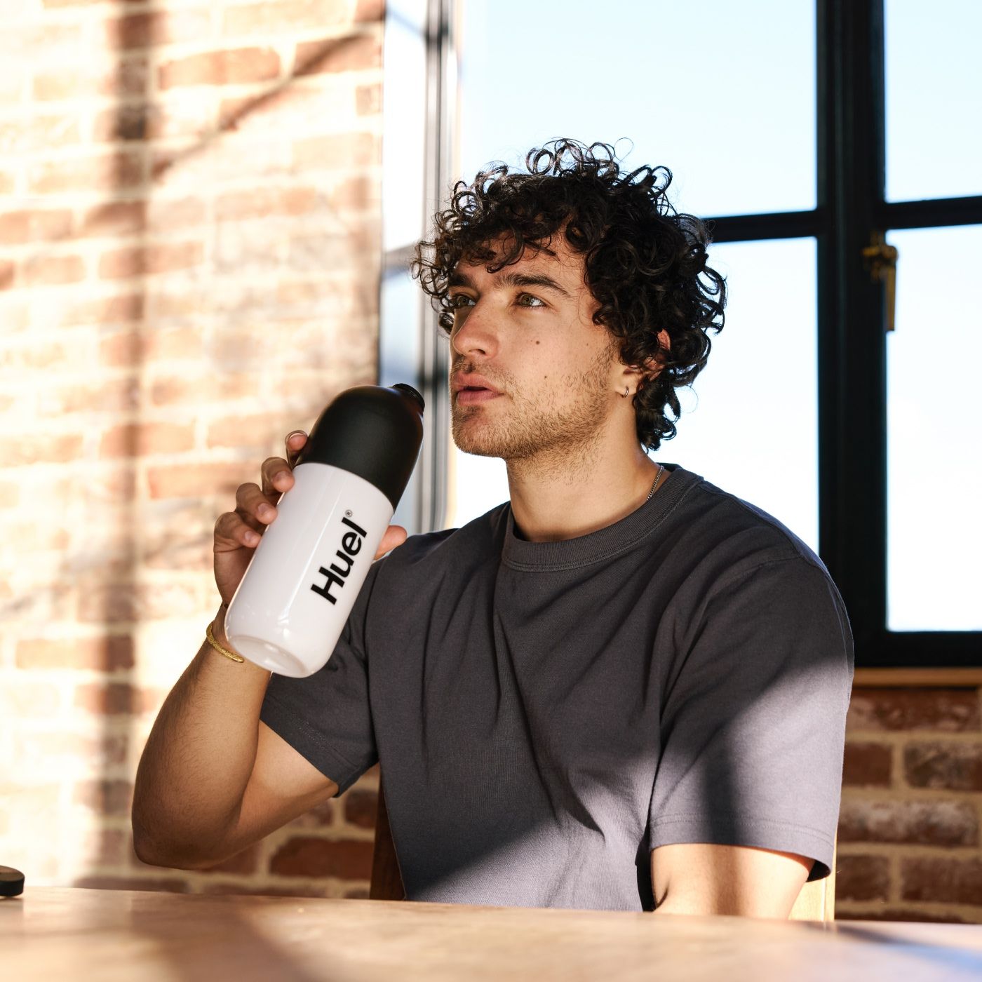 A man with curly hair is sitting indoors, sipping from a white Huel shaker with a black lid. He is seated at a wooden table in a sunlit room with large windows and exposed brick walls, enjoying a healthy meal replacement shake.
