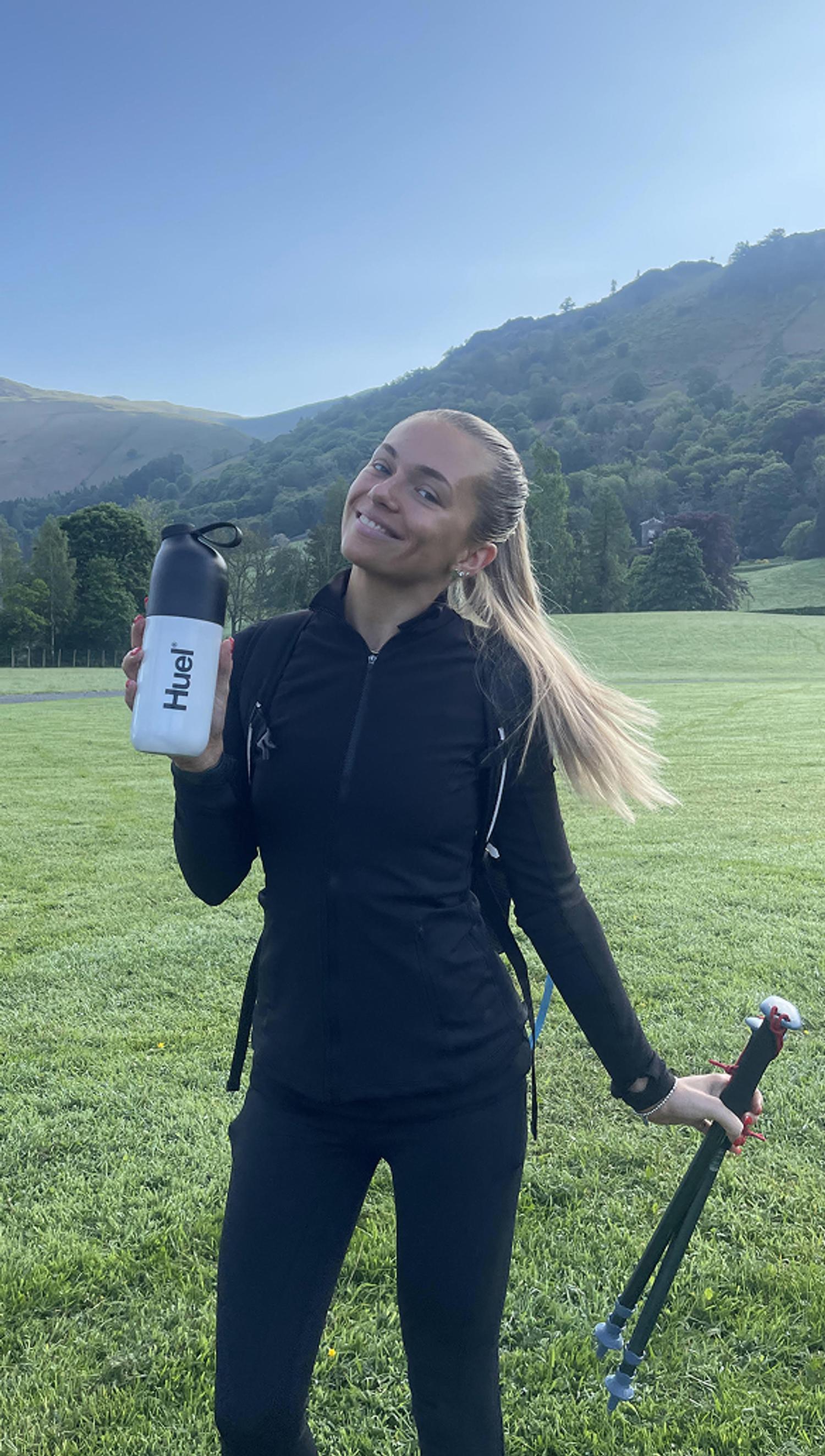 A picture of a woman holding a huel shaker in a mountainous area with walking poles in the other hand