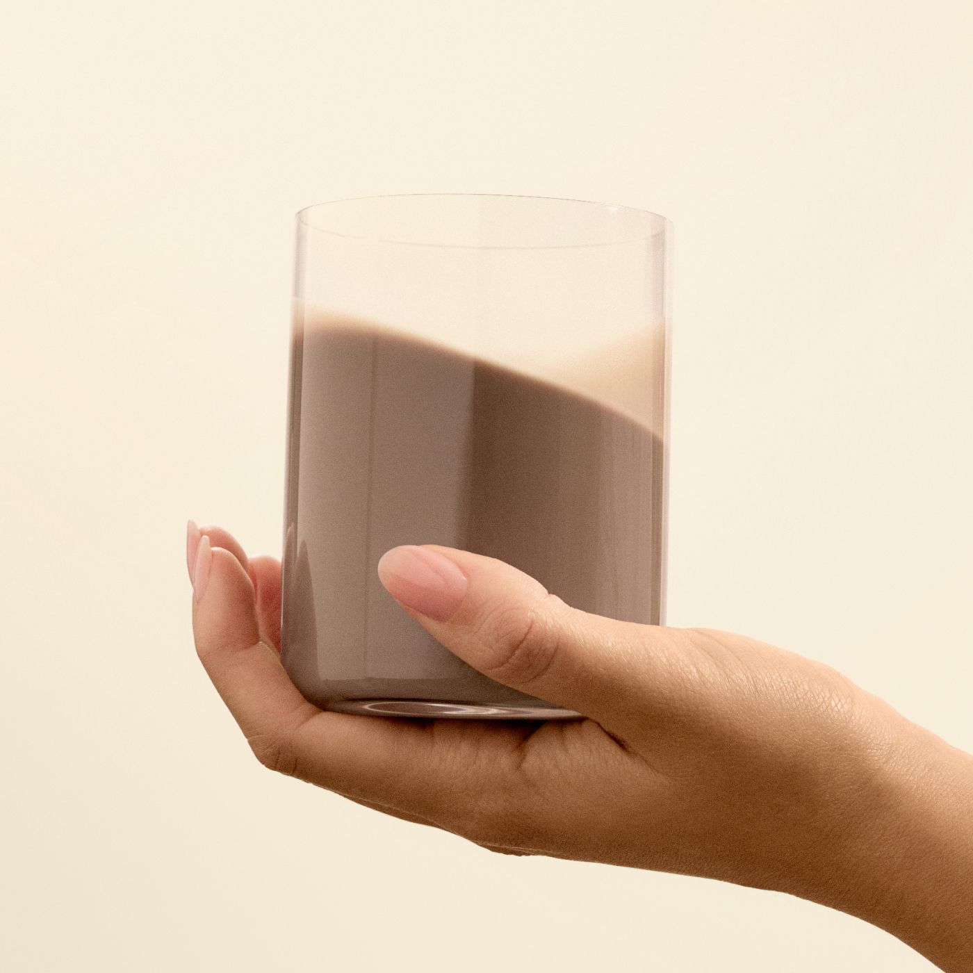 A glass being held by a hand with chocolate flavoured Huel ready-to-drink lite in it on a beige background