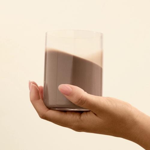 A glass being held by a hand with chocolate flavoured Huel ready-to-drink lite in it on a beige background