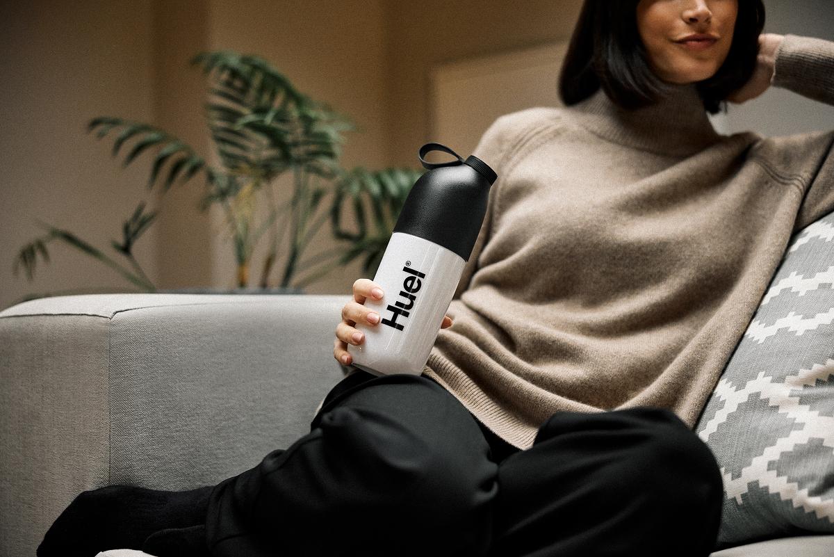 A women smiling while sat on a couch holding a huel shaker