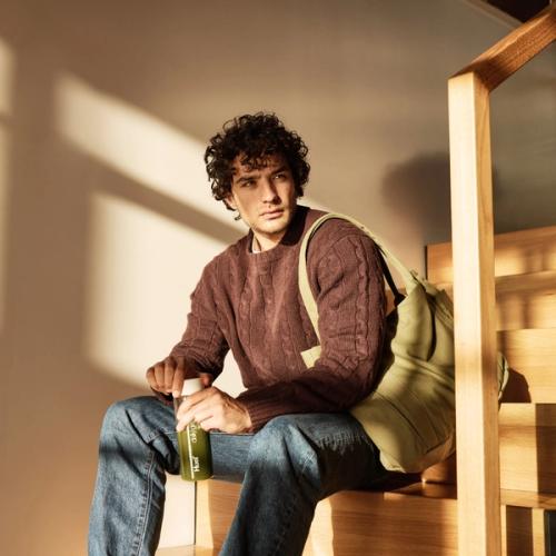 A man with curly hair sits on a wooden staircase, holding a bottle of Huel Daily Greens. He is dressed in a brown knit sweater, blue jeans, and has a green tote bag over his shoulder. Sunlight streams through a window, creating a warm, cozy atmosphere.