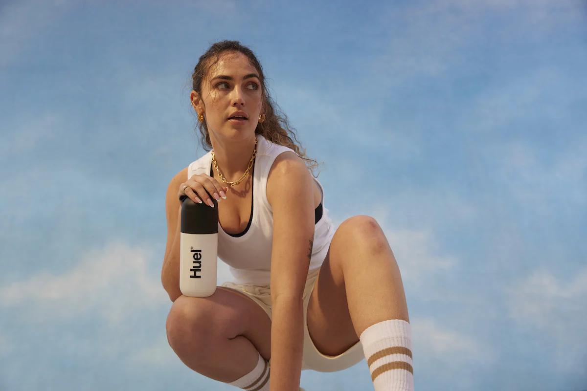 woman squatting down to rest in the sun holding a huel shaker