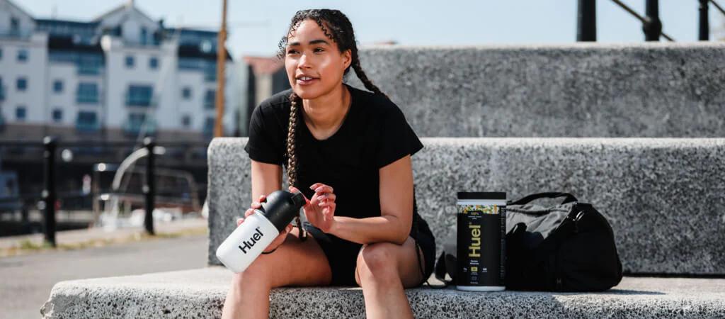 Woman sat on stone steps with Huel Complete Protein and holding a Huel shaker