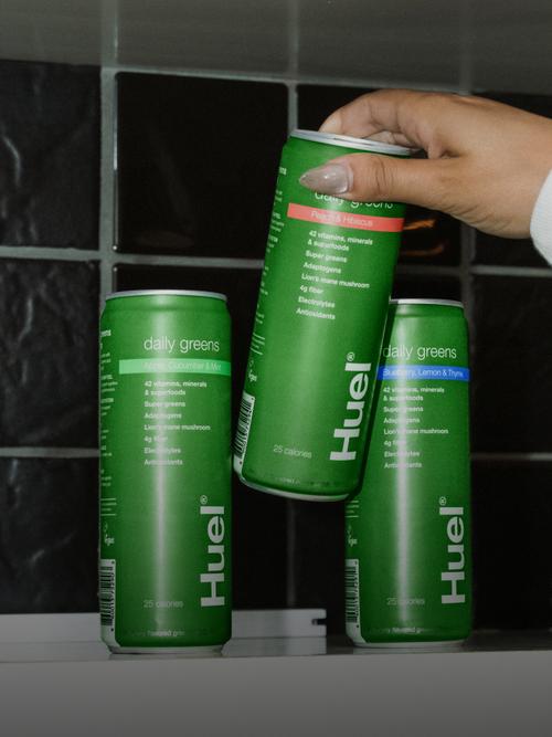 The 3 flavours of Huel Daily Greens Ready-to-drink sat on a shelf with a person lifting one of them off