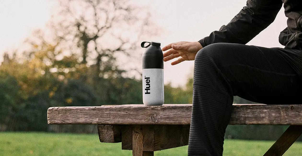 a man say on a bench outisde reaching for his huel shaker which is on the bench next to him