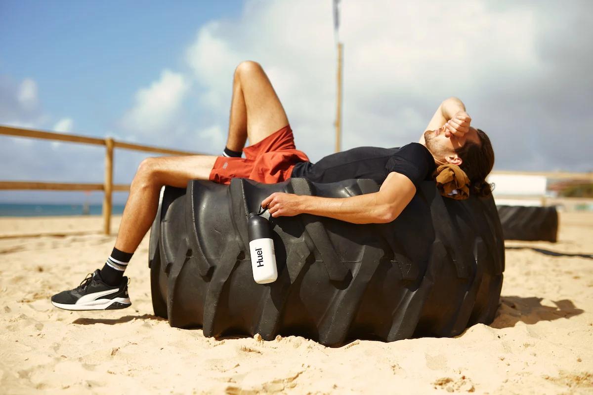 man laying on a giant tyre on the beach in the sun holding a huel shaker