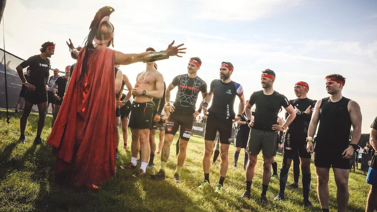 man dressed in spartan clothing and helmet facing a row of men in atheletic gear with his arms spread wide