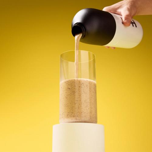 A hand holding a Huel shaker which is being poured into a glass on stand containing Huel's Black Edition Powder in Banana on a yellow background
