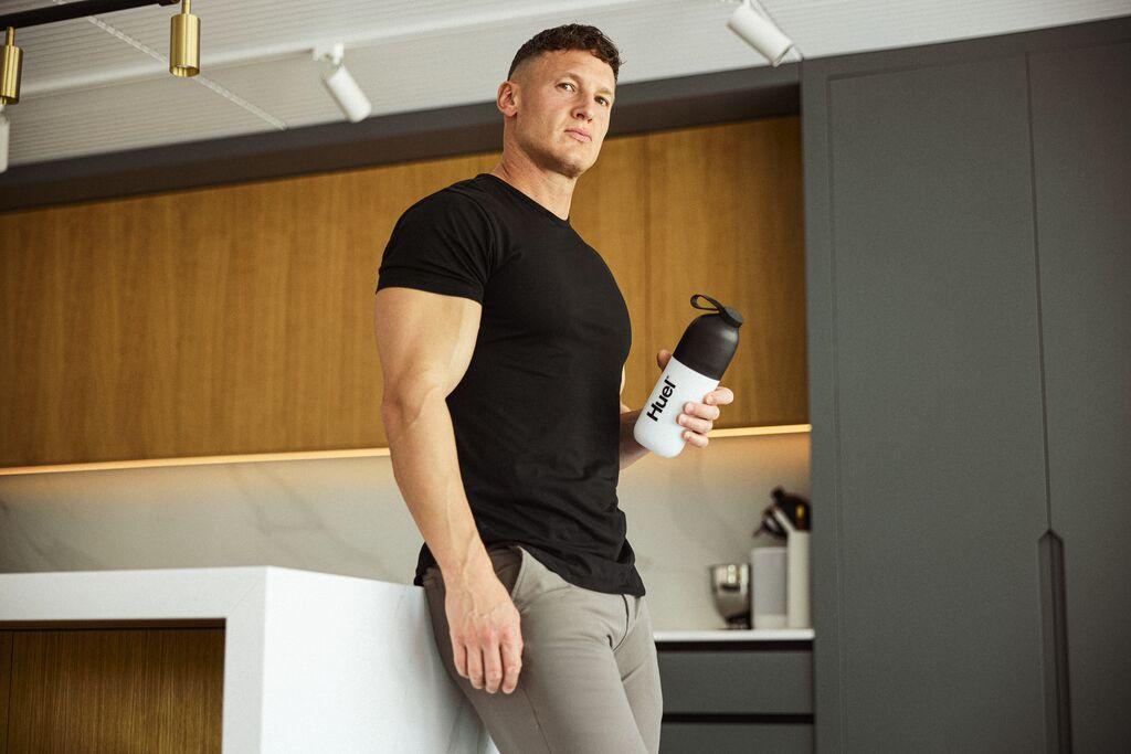 Matt Morsia leaning on a counter looking at the camera holding a Huel Shaker