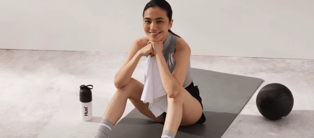 A woman sat on a yoga mat smiling next to a Huel shaker
