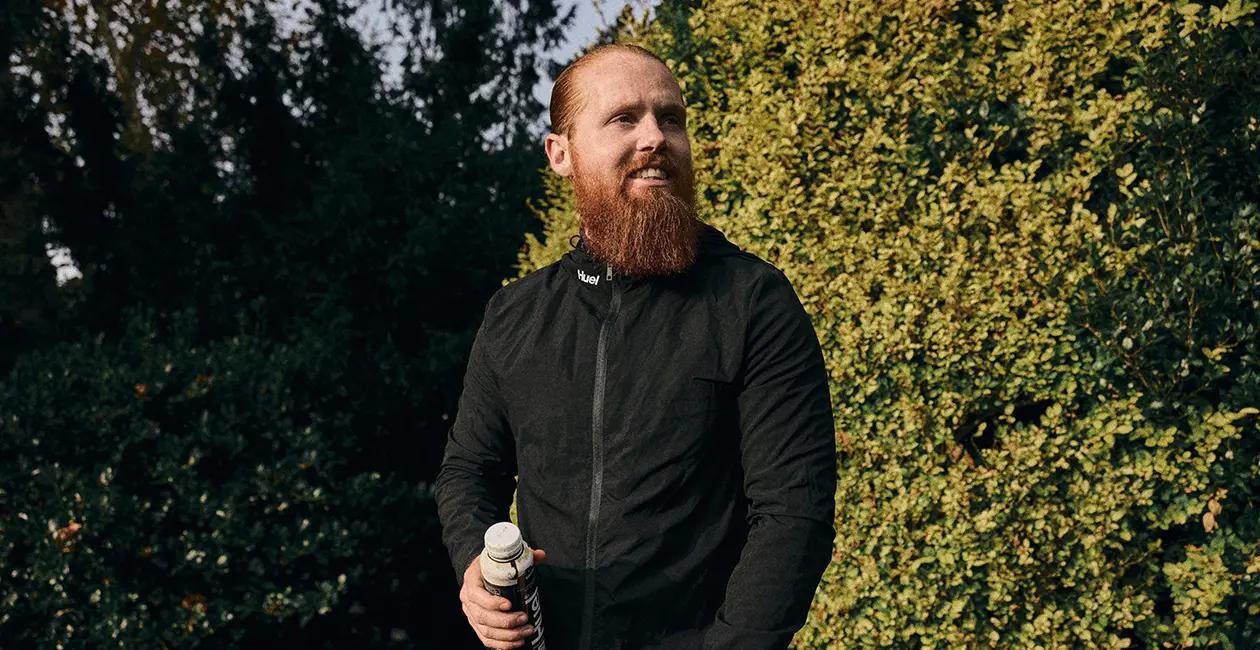 Russ Cook holding a Huel Black Edition Ready-to-drink while stood outside in front of some bushes