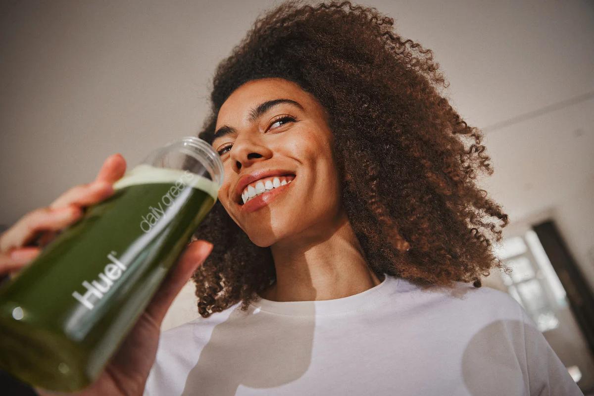 woman smiling while drinking huel daily greens from a huel bottle