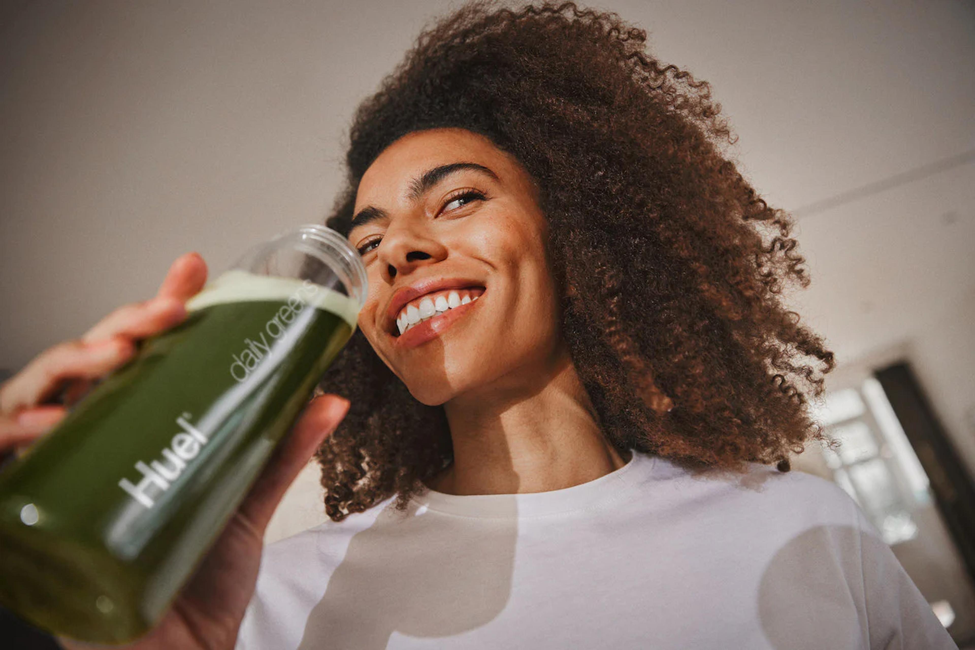 woman smiling while drinking huel daily greens from a huel bottle