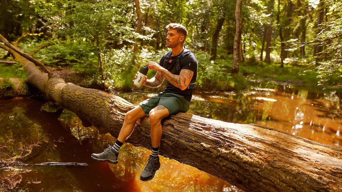Man sat on a fallen treen over a small lake holding a Huel shaker