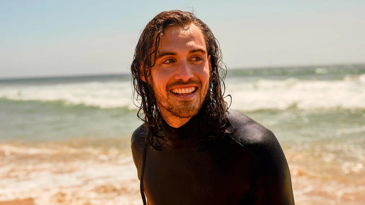 surfer on the beach in a wetsuit looking off to the right with the sea in the background