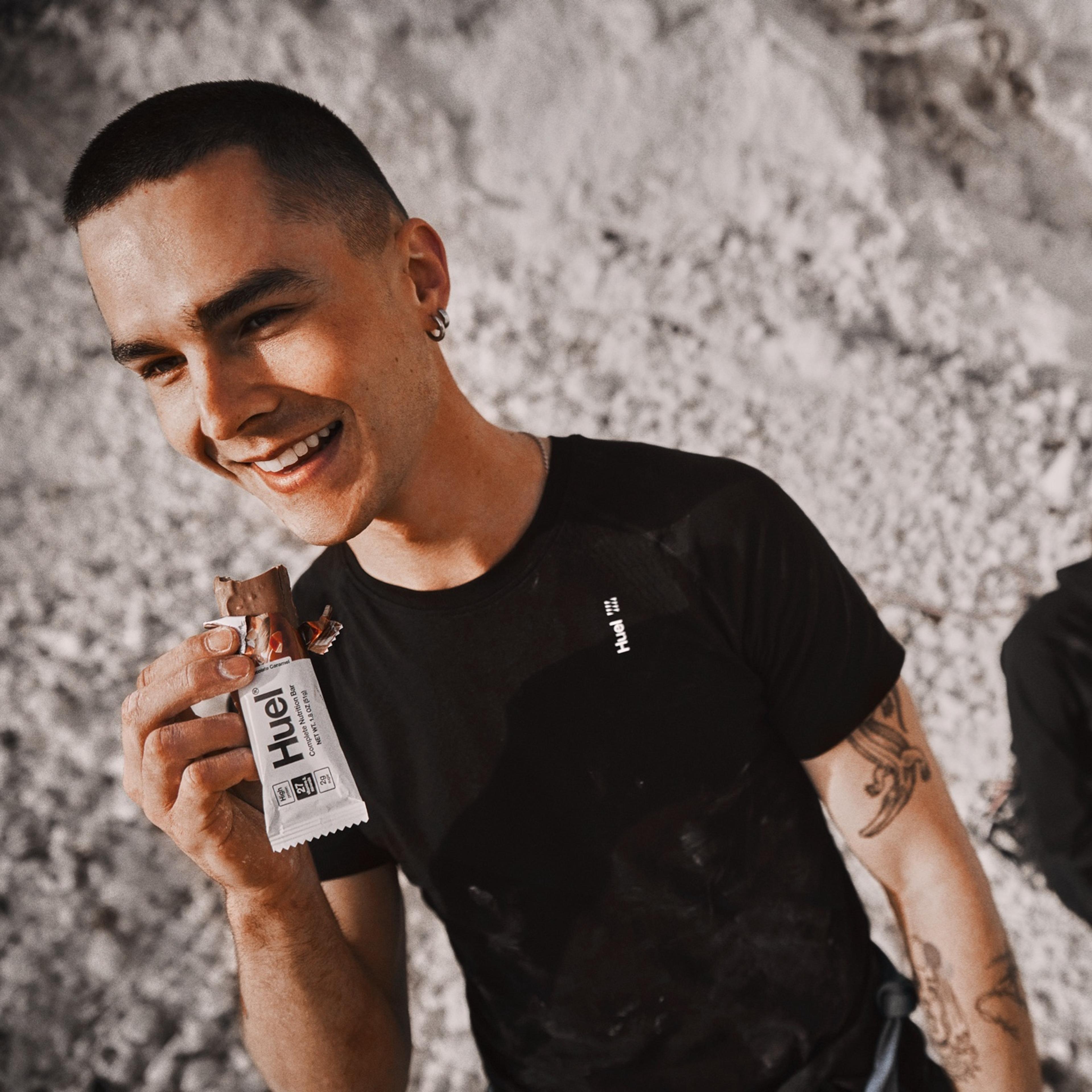A smiling man eating a Huel protein bar against a rugged mountainous background