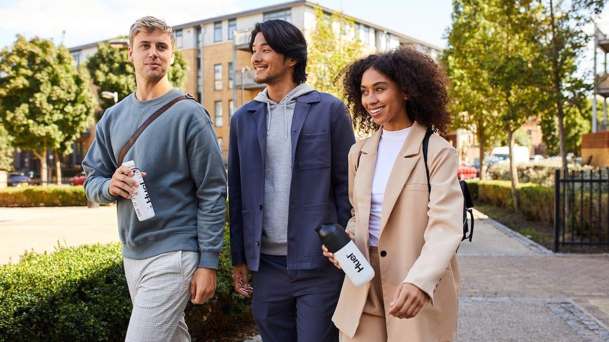 three people walking outside with trees and a building in the background one person holding a Huel RTD and another holding a Huel shaker