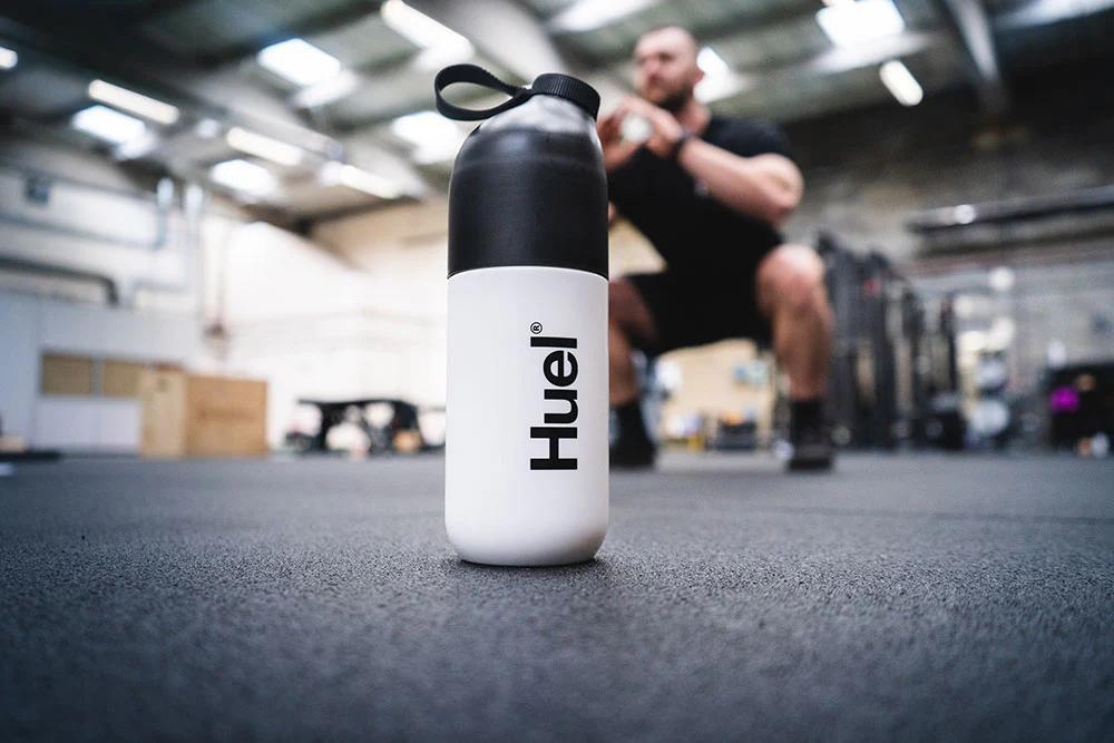 a huel shaker in the foreground on the floor of a gym with a man blurred in the background doing squats