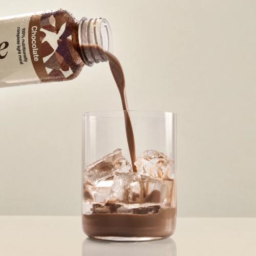 Huel chocolate pouring from a bottle of Huel Ready-to-drink lite into a glass with ice on a beige background