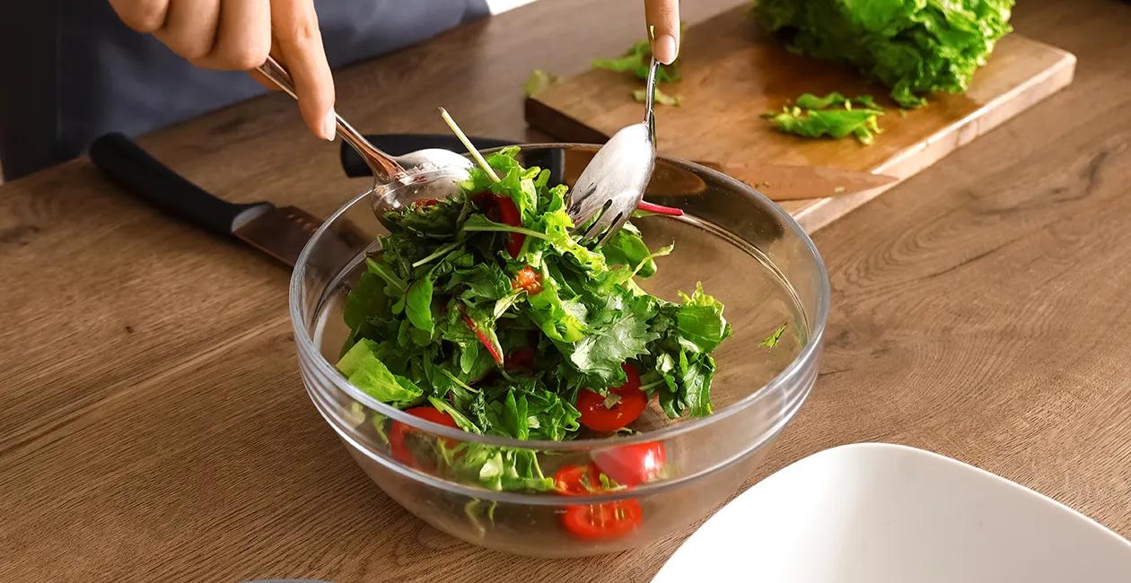 person tossing a leafy salad with a chopping board knife and lettuce in the background
