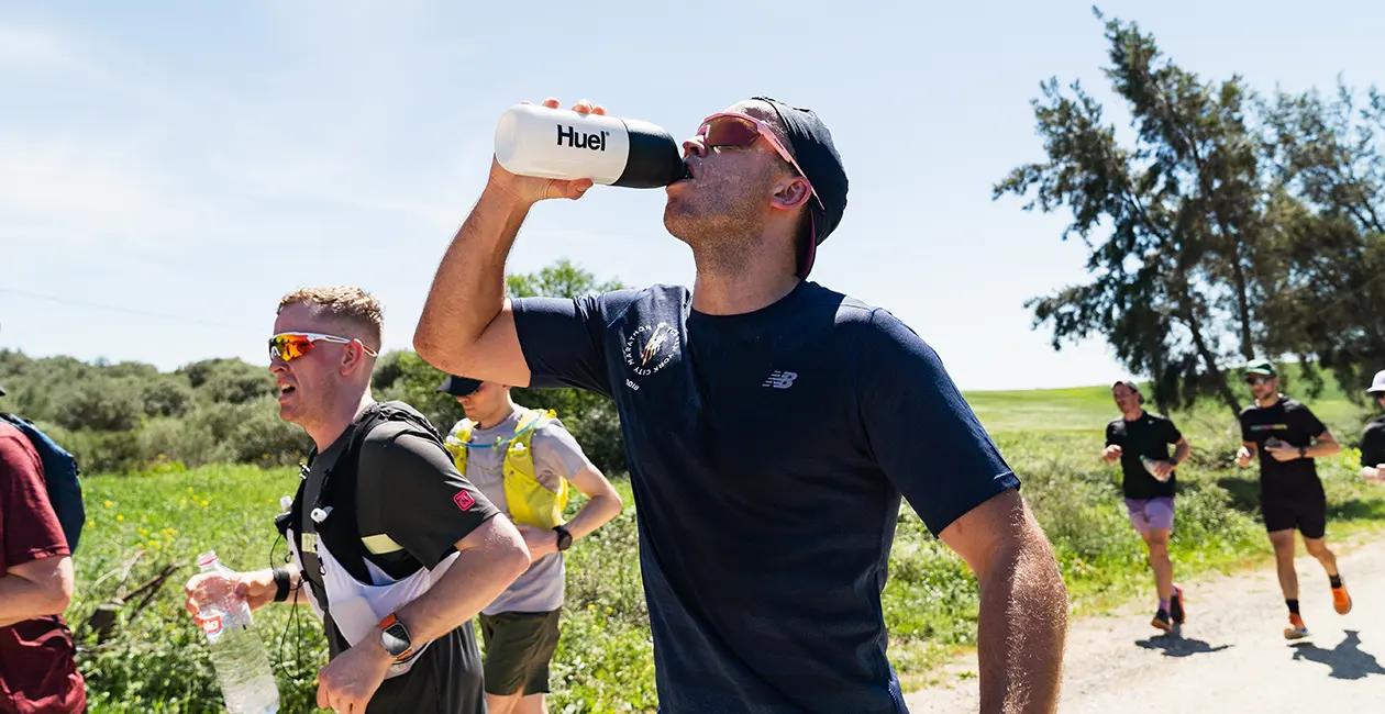 men running along a trail outside in the sun one of them drinking from a Huel shaker