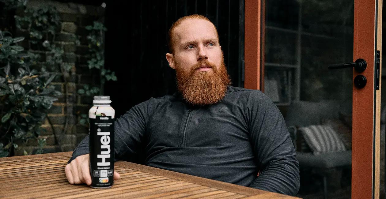 Russ Cook sat at a table looking off into the distance with a Huel Black Edition Ready-to-drink on the table