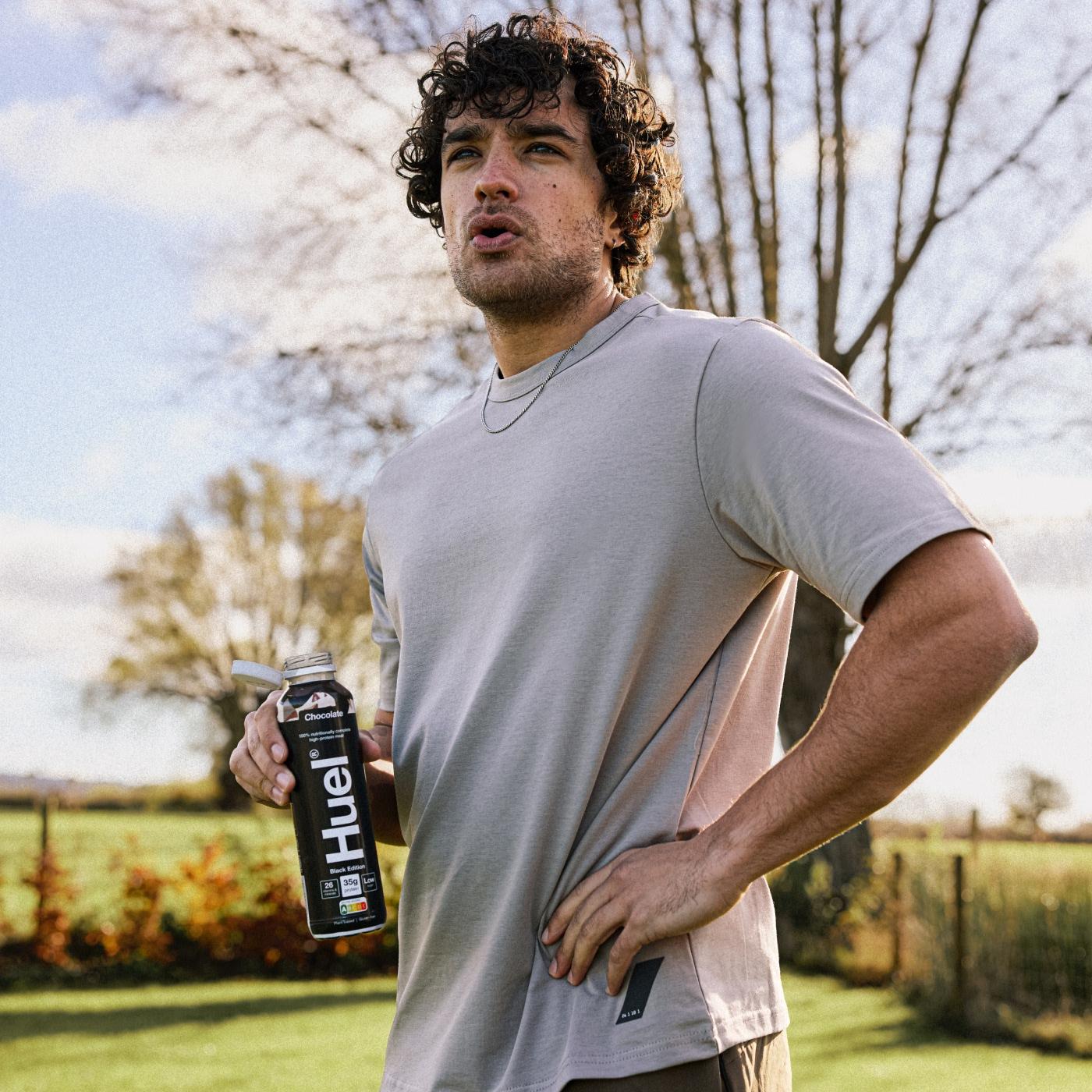 A man with curly hair stands outdoors holding a Huel Chocolate Ready-to-Drink bottle. He wears a beige T-shirt with one hand on his hip. The background shows a grassy field, trees, and a clear sky, creating a calm countryside setting.