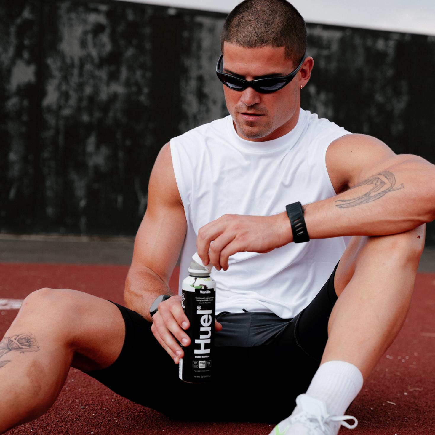 A man wearing a sleeveless white tank and black shorts is sitting on ground unscrewing the top off a Black Edition Ready-to-drink