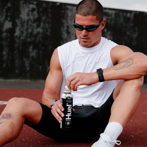 A man wearing a sleeveless white tank and black shorts is sitting on ground unscrewing the top off a Black Edition Ready-to-drink