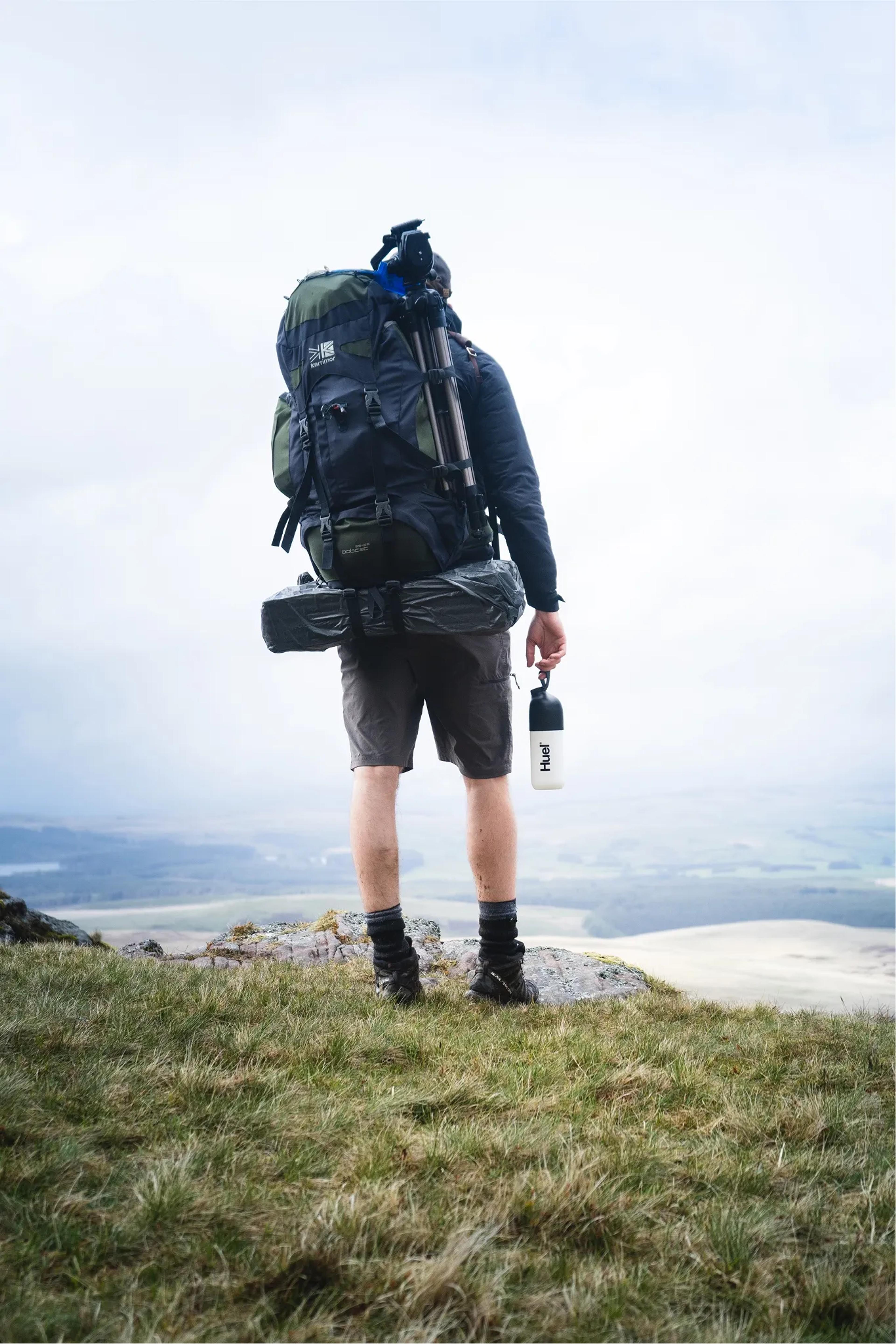 man stood at top of a hill overlooking a valley holding a Huel shaker