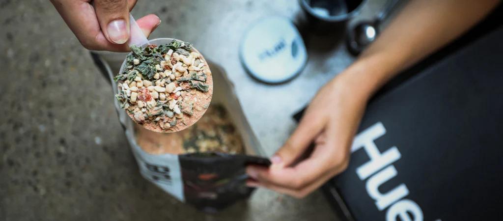 A single scoop of Huel Instant Hot Meal being held above a pouch of the Huel Hot and Savoury
