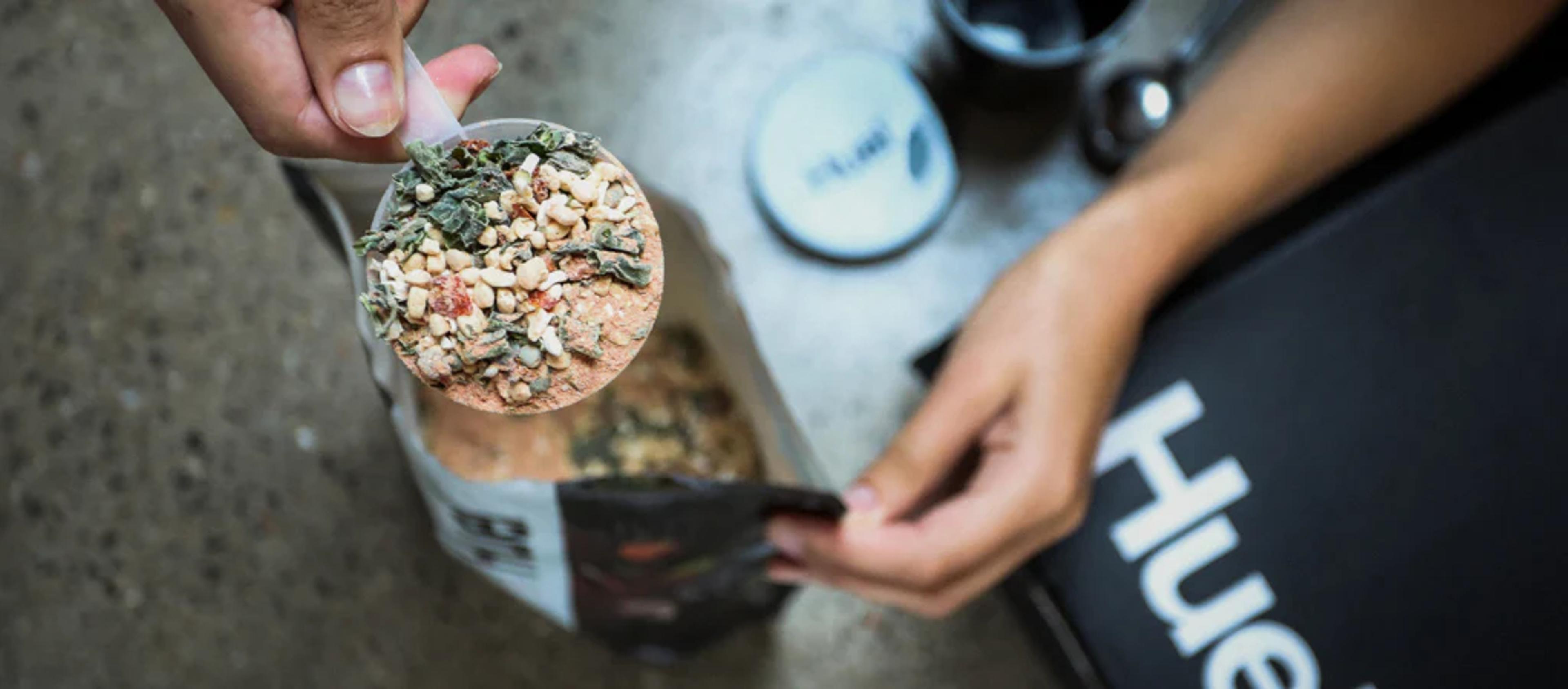 A single scoop of Huel Instant Hot Meal being held above a pouch of the Huel Hot and Savoury