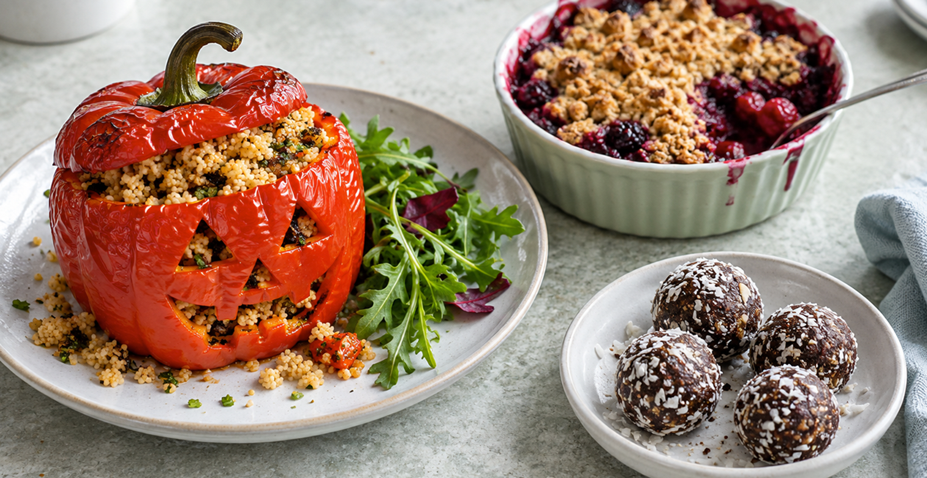 A roasted pumpkin with a carved face full of couscous with a side of salad. There is also a crumble dessert and protein balls on other dishes