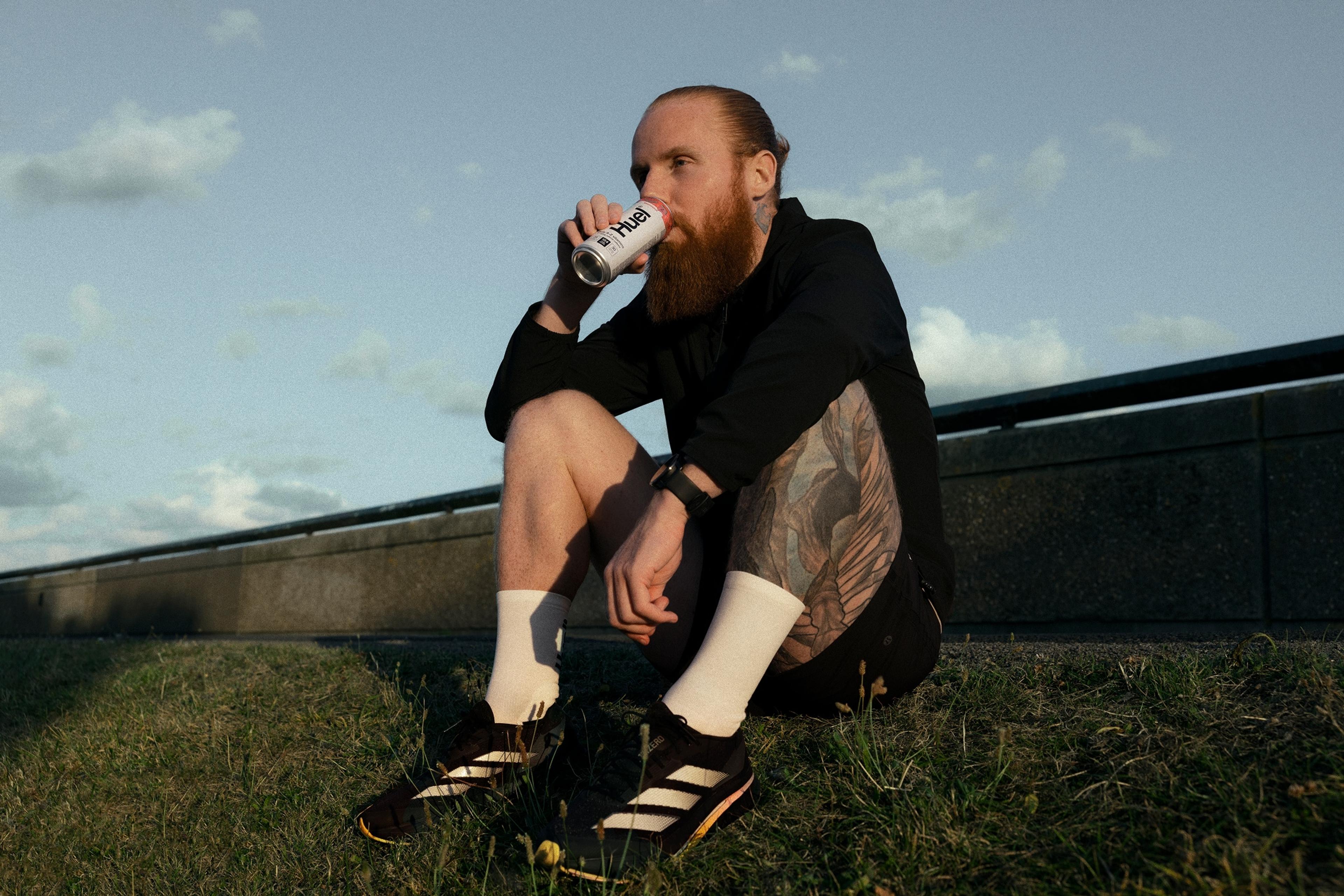 Russ Cook sat on the ground outside drinking a Huel Daily A to Z vitamins drink
