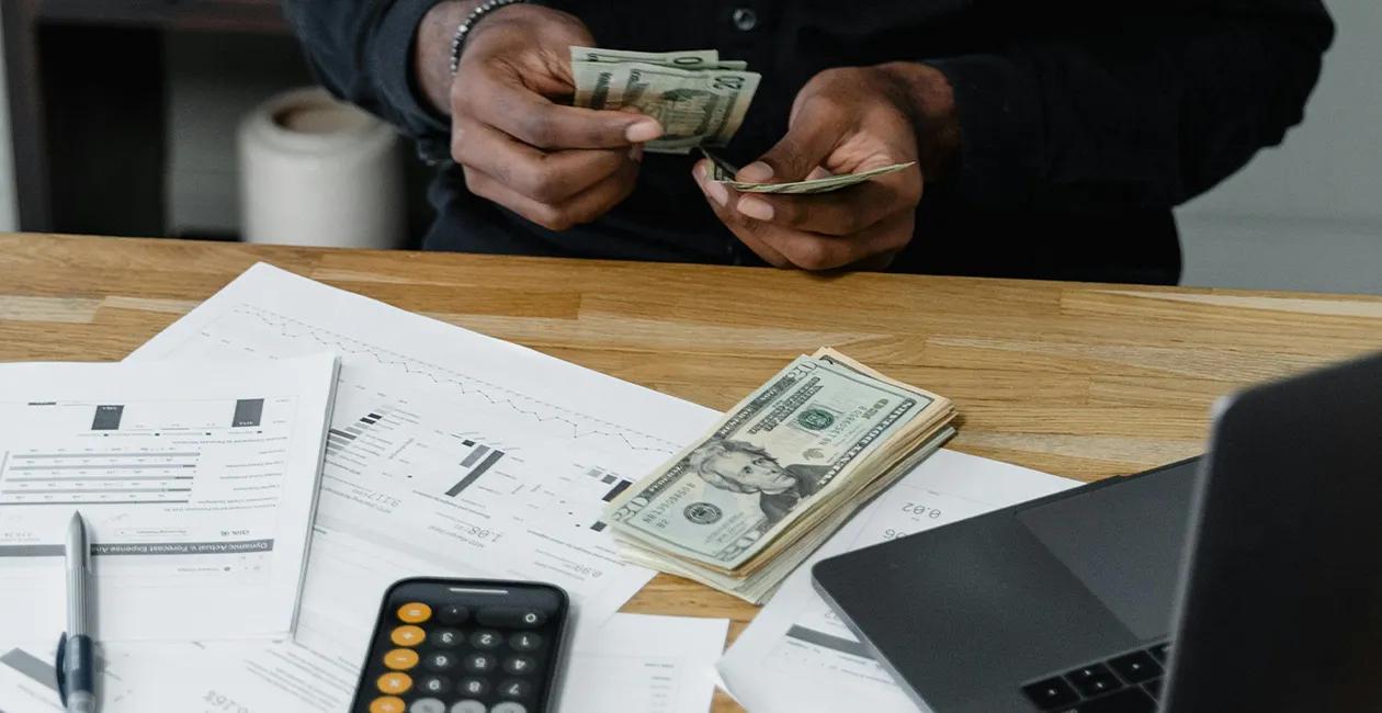 person sat at a desk counting money with their laptop bills and calculator on the desk
