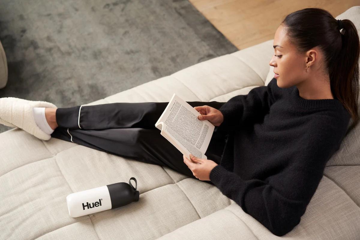 woman sat on sofa with her feet up reading a book and with a huel shaker on the sofa next to her
