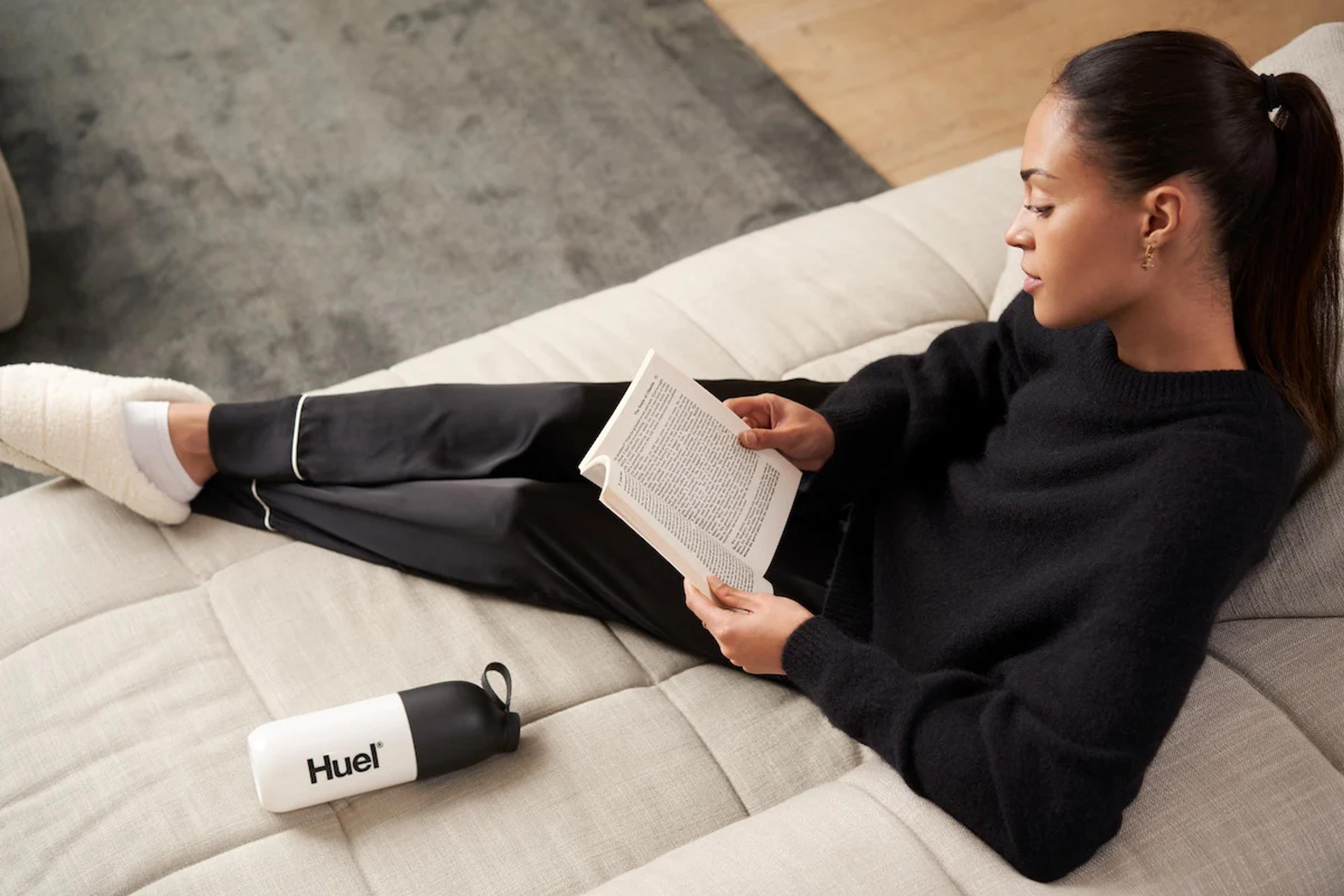 woman sat on sofa with her feet up reading a book and with a huel shaker on the sofa next to her