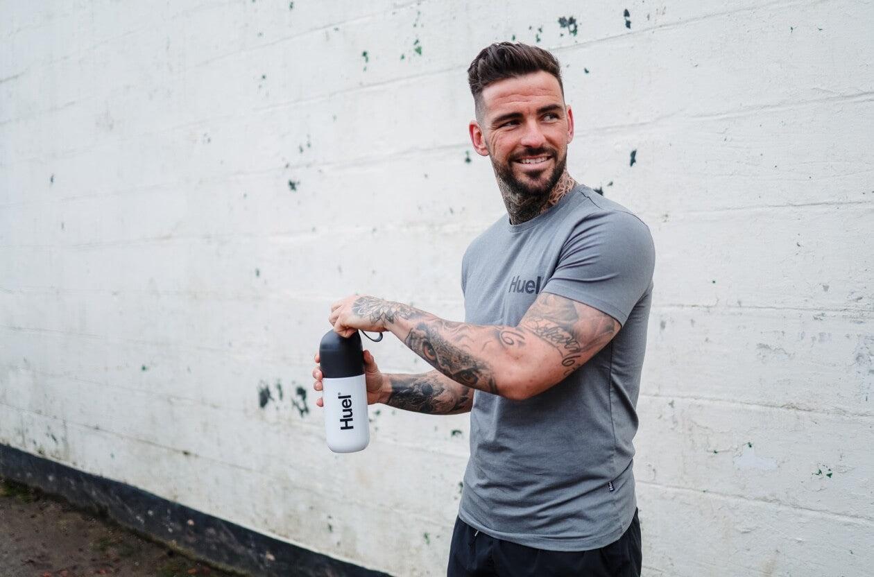 A Man holding a Huel shaker in a grey Huel branded shirt in front of a white brick background