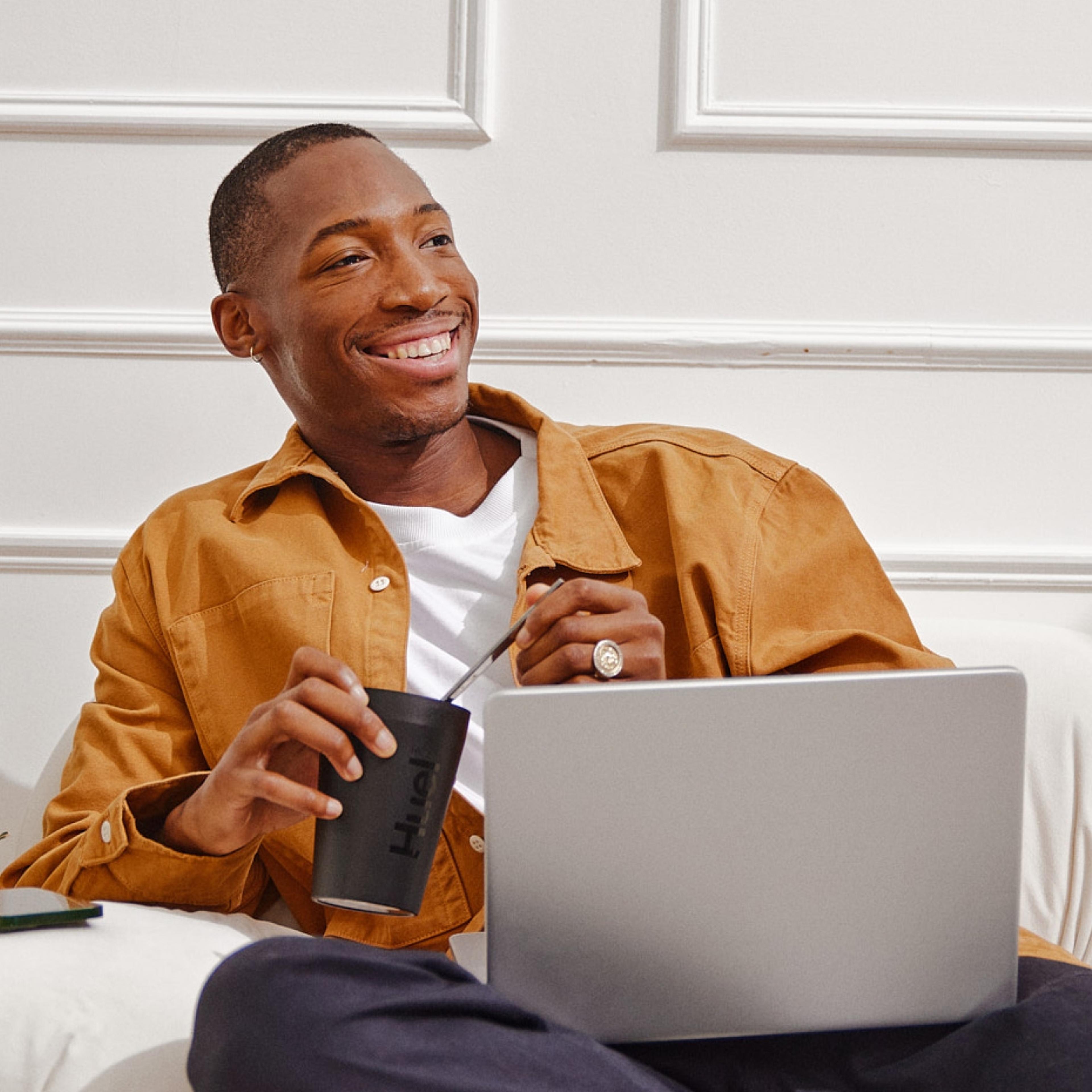 A smiling man in a mustard-colored jacket sits on a couch with a laptop on his lap. He is holding a black Huel cup and stirring its contents, creating a relaxed, work-from-home vibe in a bright, stylish room with white walls.