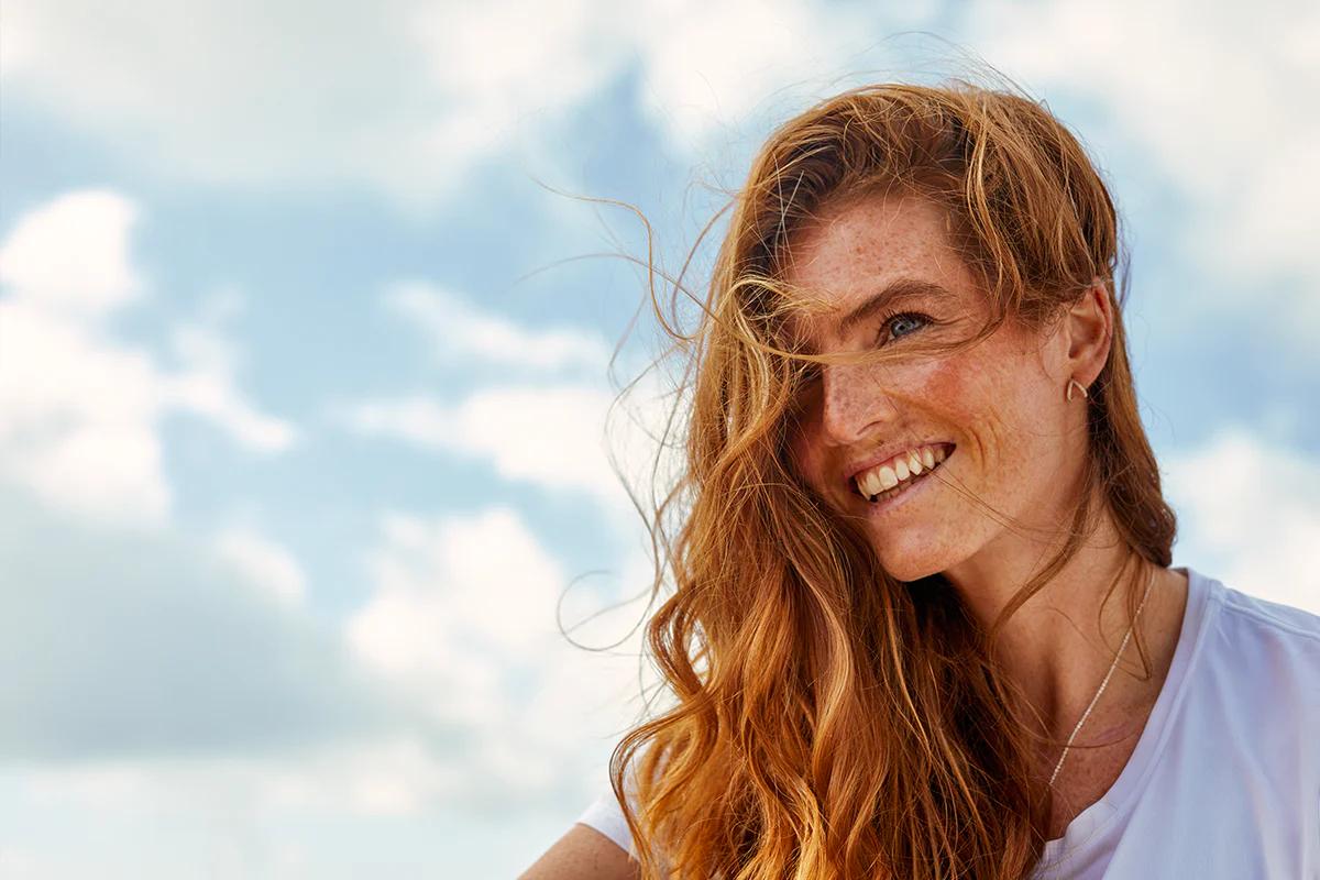 woman with ginger hair and freckles smiling while outside with clouds in the background