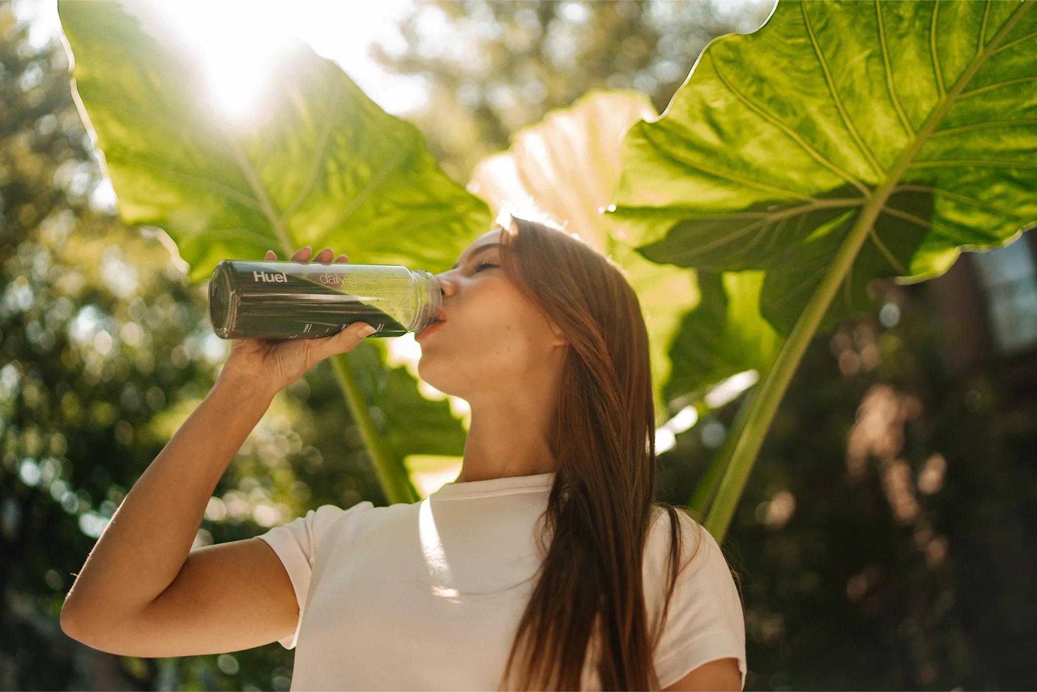 woman stood outside in front of leafy plants drinking Huel Daily Greens