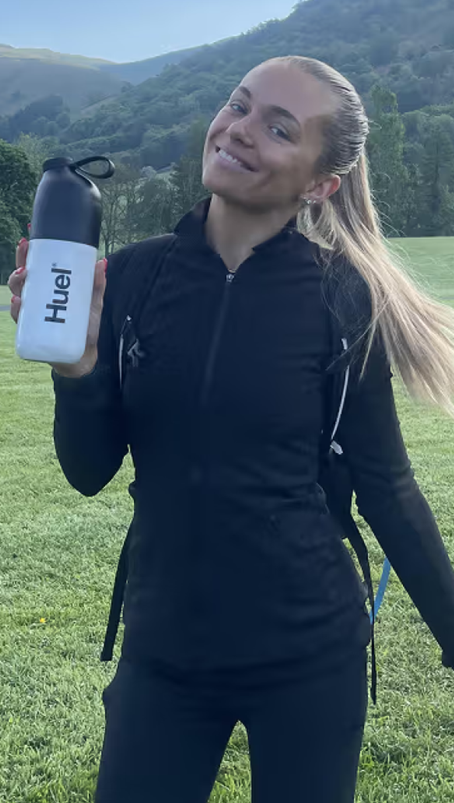 A picture of a woman holding a huel shaker in a mountainous area with walking poles in the other hand