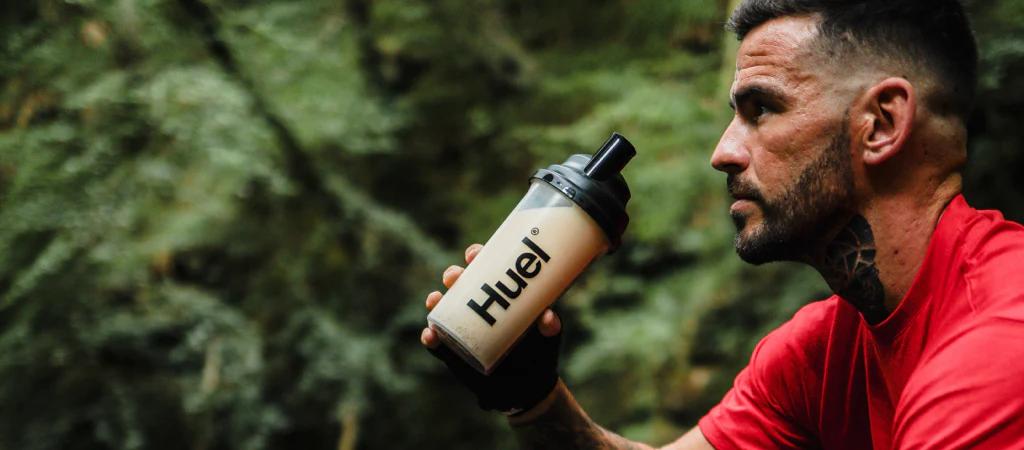 man drinking from a huel shaker in the woods