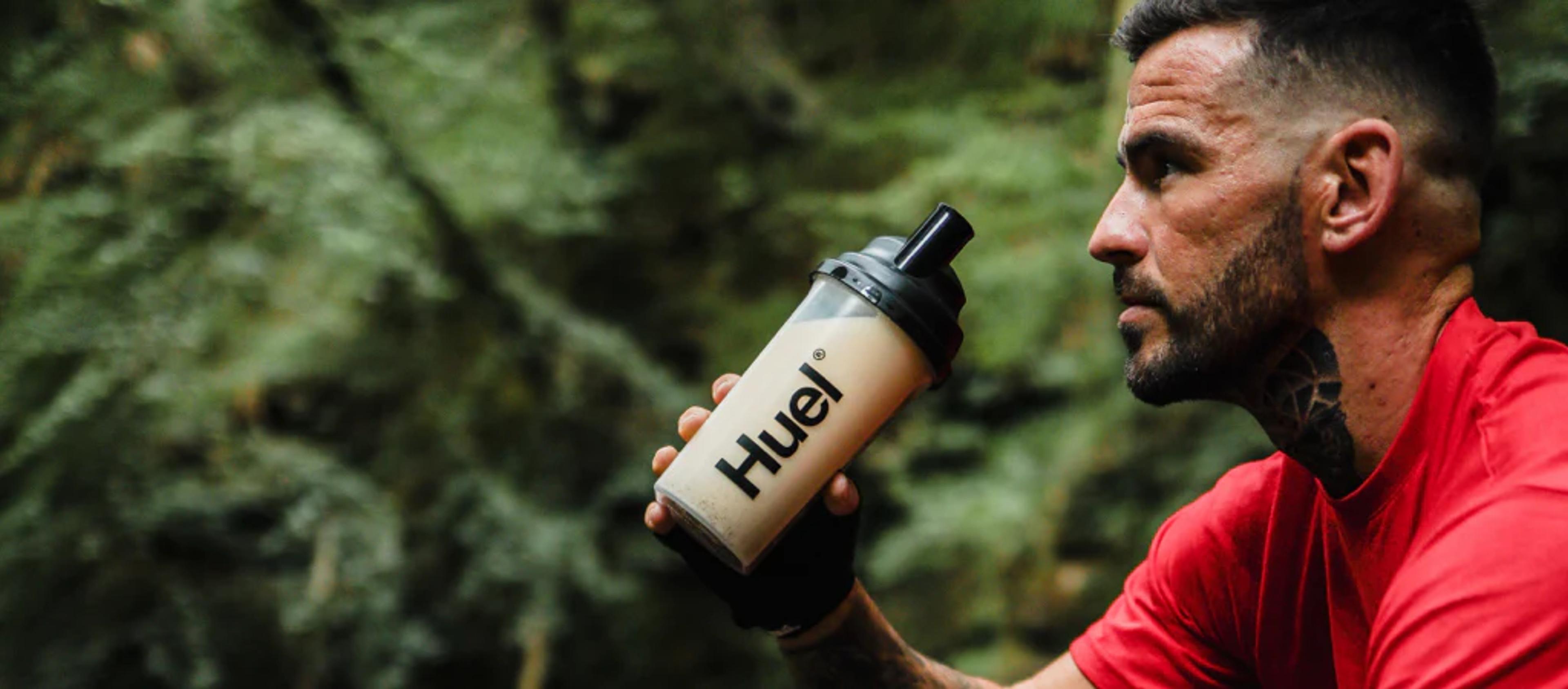 man drinking from a huel shaker in the woods