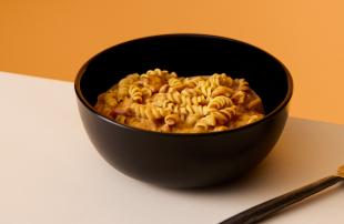 A bowl of Cajun Pasta Huel Hot & Savoury, on a table, set against a yellow background