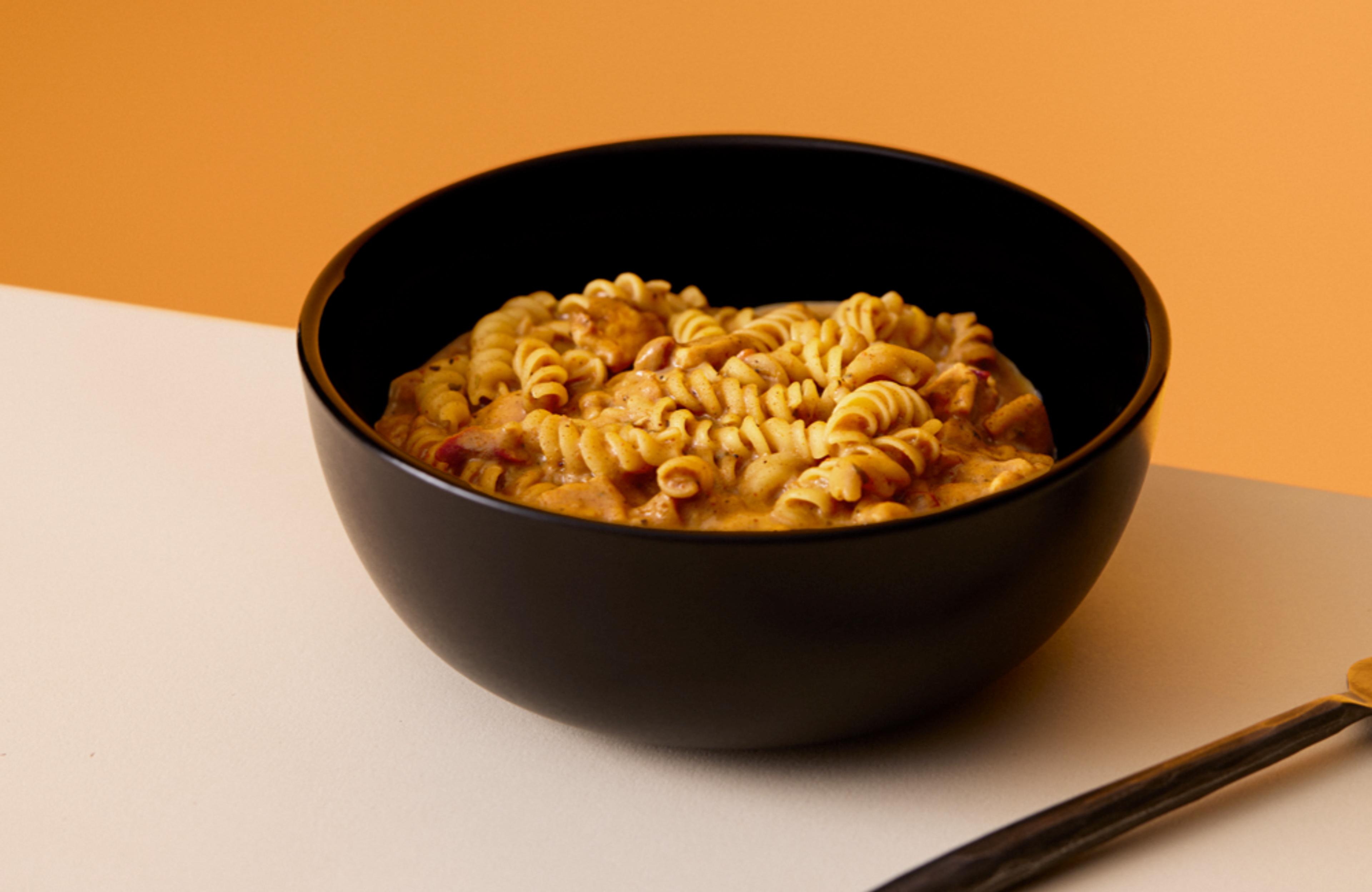 A bowl of Cajun Pasta Huel Hot & Savoury, on a table, set against a yellow background