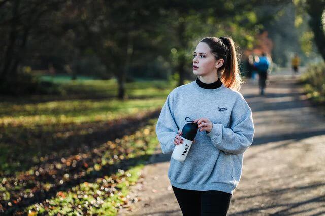 woman walking along a path in a park holding a huel shaker and looking off into hte distance