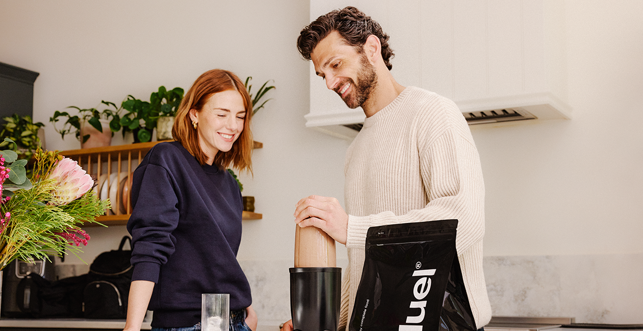 A man and woman standing over a counter with a bag of Huel powder and a blender