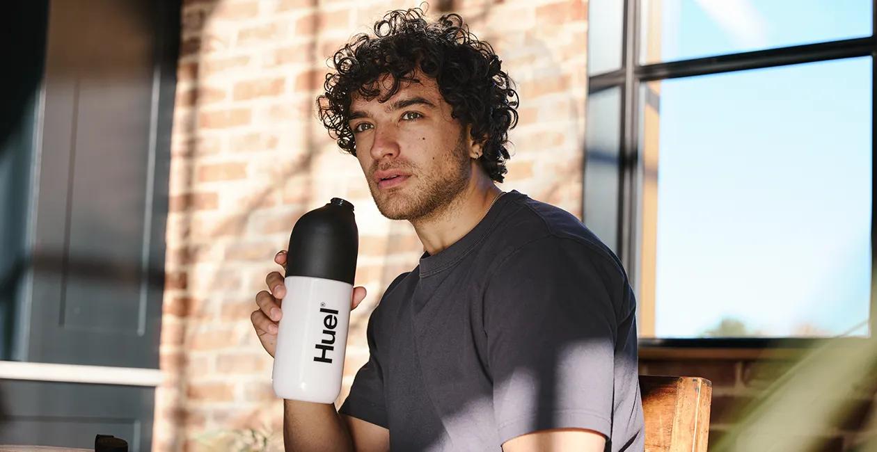 man sat inside infront of a window holding a Huel shaker looking longingly into the distance